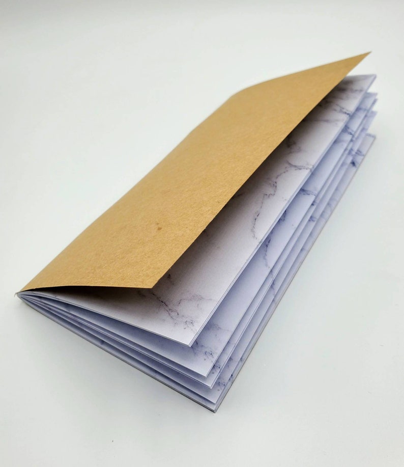 May include: A brown paper covered notebook with white pages featuring a marble pattern. The notebook is open to reveal the pages.
