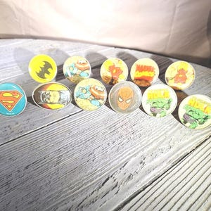 May include: A collection of round, vintage superhero pins featuring characters like Superman, Batman, Captain America, Spider-Man, The Hulk, and The Flash. The pins have a silver border and are arranged on a gray wooden surface.