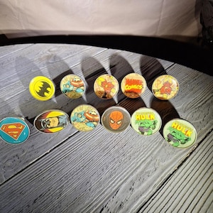 May include: A collection of comic book character pins, including Batman, Superman, Captain America, Spider-Man, and the Hulk. The pins are round and oval, with colorful graphics and silver-colored metal edges. The Marvel Comics logo is also visible.