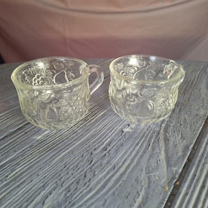 Peut inclure: Deux tasses en verre transparent avec un motif de fruits. L'une des tasses a une anse, l'autre n'en a pas. Les tasses sont posées sur une surface en bois grise. Les tasses sont probablement destinées à servir des boissons.