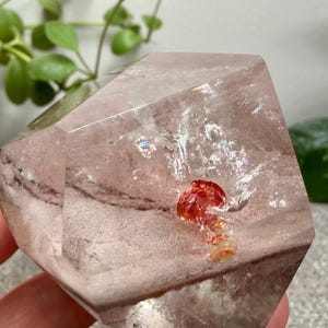 May include: A faceted, translucent quartz crystal with a pale pinkish-brown hue. The interior features a distinct, vibrant red mineral inclusion and a small, shimmering iridescent patch, showcasing natural internal fractures and geometric angles.