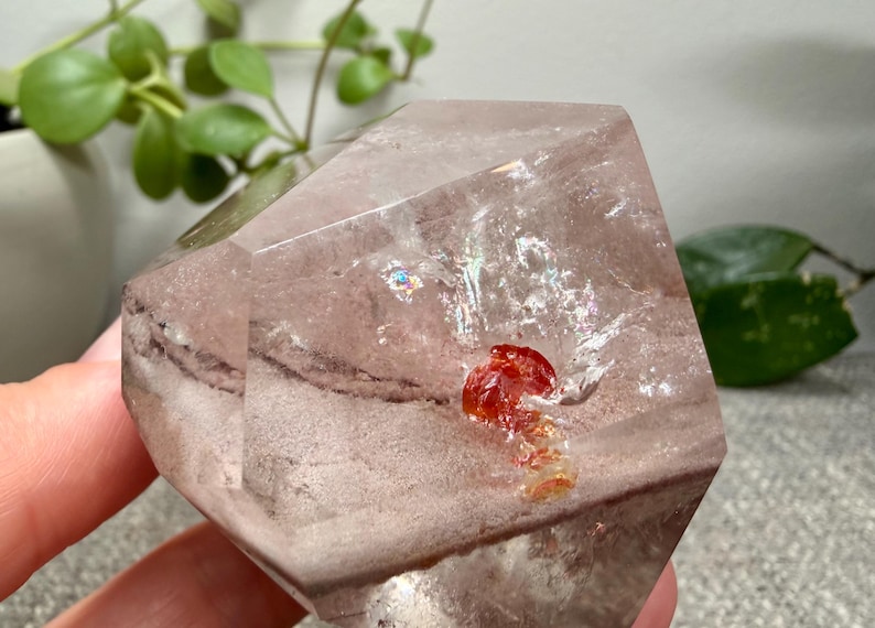 May include: A faceted, translucent quartz crystal with a pale pinkish-brown hue. The interior features a distinct, vibrant red mineral inclusion and a small, shimmering iridescent patch, showcasing natural internal fractures and geometric angles.