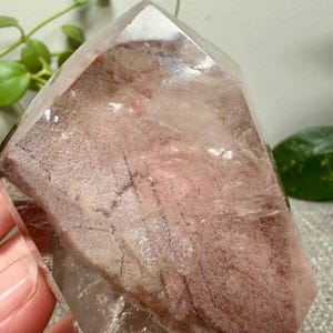 May include: A clear, pointed quartz crystal specimen featuring a prominent internal phantom inclusion. The interior displays a dusty, reddish-brown, textured layer with distinct geometric patterns and fractures visible through the transparent outer surface.