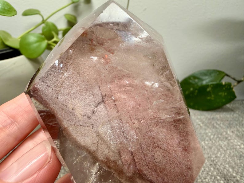 May include: A clear, pointed quartz crystal specimen featuring a prominent internal phantom inclusion. The interior displays a dusty, reddish-brown, textured layer with distinct geometric patterns and fractures visible through the transparent outer surface.