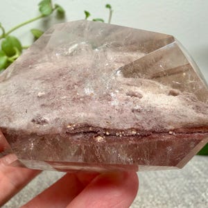 May include: A polished, semi-translucent quartz crystal specimen featuring a prominent internal layer of reddish-brown, earthy mineral inclusions. The crystal has a smooth, faceted surface with clear edges and a distinct, textured, matte-finish core.