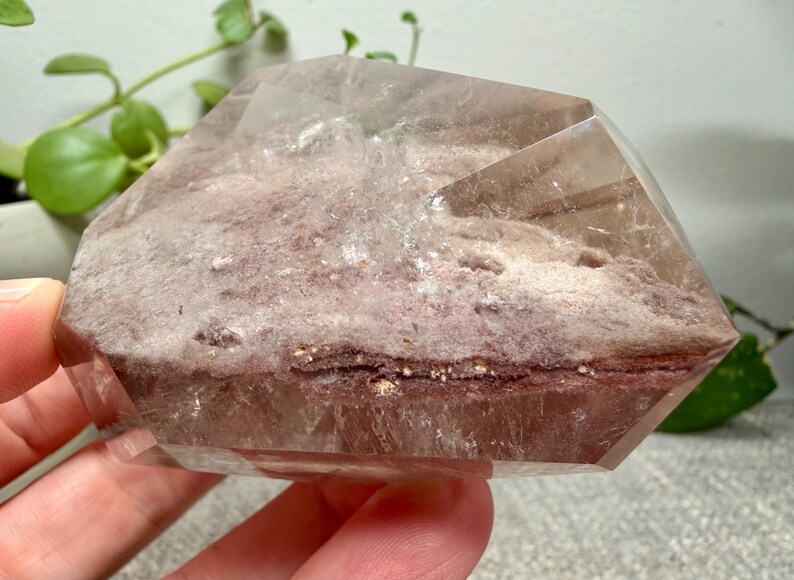 May include: A polished, semi-translucent quartz crystal specimen featuring a prominent internal layer of reddish-brown, earthy mineral inclusions. The crystal has a smooth, faceted surface with clear edges and a distinct, textured, matte-finish core.