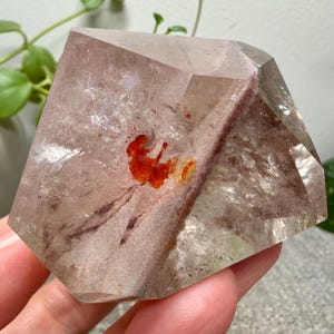 May include: A polished, multifaceted clear quartz crystal featuring internal inclusions of reddish-brown mineral deposits and a distinct, vibrant orange-red patch near the center, showcasing natural geometric facets and internal clarity.
