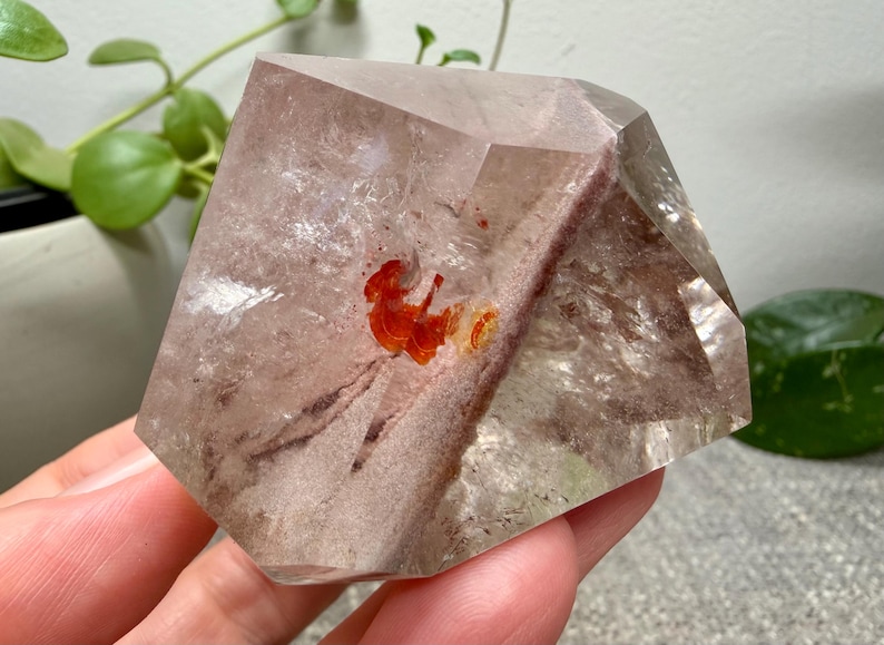 May include: A polished, multifaceted clear quartz crystal featuring internal inclusions of reddish-brown mineral deposits and a distinct, vibrant orange-red patch near the center, showcasing natural geometric facets and internal clarity.