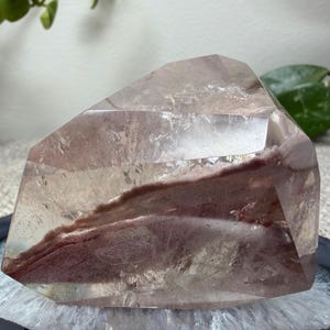 May include: A polished, clear quartz crystal specimen featuring a prominent, jagged internal inclusion of reddish-brown mineral matter. The crystal has multiple flat, faceted surfaces and a translucent appearance, revealing the deep earthy color.