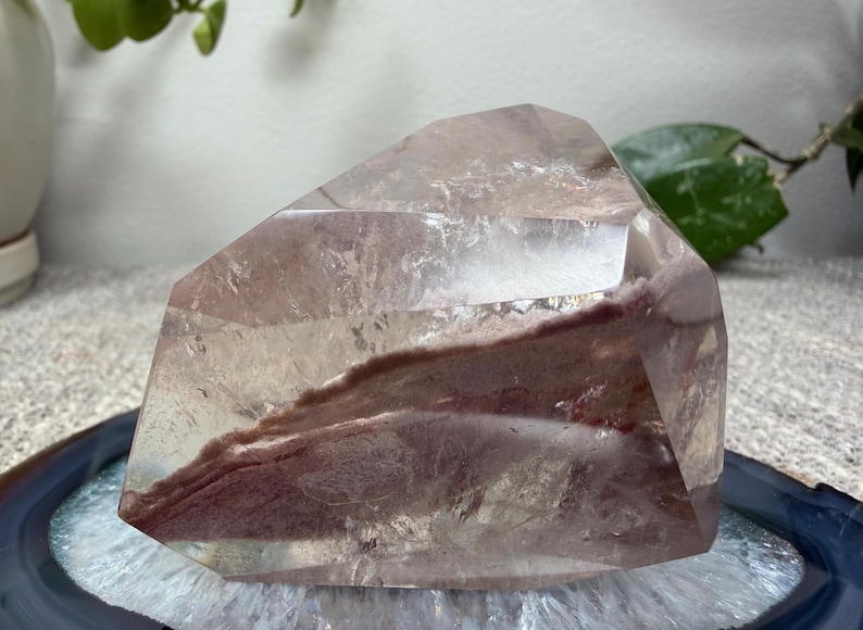 May include: A polished, clear quartz crystal specimen featuring a prominent, jagged internal inclusion of reddish-brown mineral matter. The crystal has multiple flat, faceted surfaces and a translucent appearance, revealing the deep earthy color.