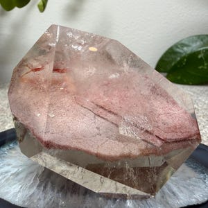 May include: A clear quartz crystal specimen featuring a prominent internal layer of dusty rose-colored mineral inclusions. The crystal has a polished, multifaceted surface with visible natural fractures and a distinct, flat, reddish-pink phantom formation.