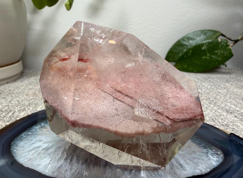 May include: A clear quartz crystal specimen featuring a prominent internal layer of dusty rose-colored mineral inclusions. The crystal has a polished, multifaceted surface with visible natural fractures and a distinct, flat, reddish-pink phantom formation.