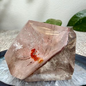 May include: A polished, multifaceted quartz crystal specimen featuring a translucent, smoky brown body with a prominent, opaque, dusty rose-colored inclusion and a small, vibrant, bright orange mineral deposit embedded within the center.