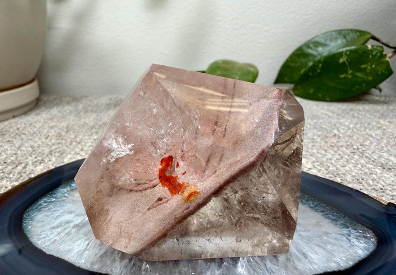 May include: A polished, multifaceted quartz crystal specimen featuring a translucent, smoky brown body with a prominent, opaque, dusty rose-colored inclusion and a small, vibrant, bright orange mineral deposit embedded within the center.