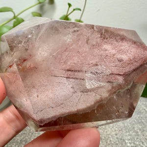 May include: A polished, multifaceted quartz crystal specimen featuring a prominent, textured reddish-brown inclusion layer within its clear, transparent body. The stone displays natural internal fractures and geometric facets throughout its structure.