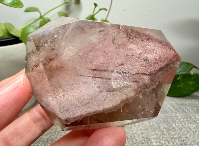 May include: A polished, multifaceted quartz crystal specimen featuring a prominent, textured reddish-brown inclusion layer within its clear, transparent body. The stone displays natural internal fractures and geometric facets throughout its structure.