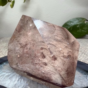 May include: A polished, multifaceted crystal point featuring a dusty rose and mauve color palette with internal cloudy inclusions and a smooth, reflective surface. The stone has a distinct geometric shape with sharp edges and a pointed top section.