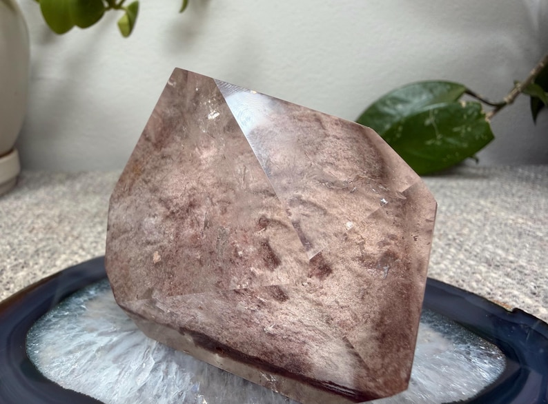 May include: A polished, multifaceted crystal point featuring a dusty rose and mauve color palette with internal cloudy inclusions and a smooth, reflective surface. The stone has a distinct geometric shape with sharp edges and a pointed top section.