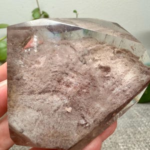 May include: A polished, faceted quartz crystal specimen featuring a clear, transparent outer layer and a textured, dusty rose-colored interior inclusion. The stone has a flat, geometric top surface and a wide, irregular base with visible mineral deposits.