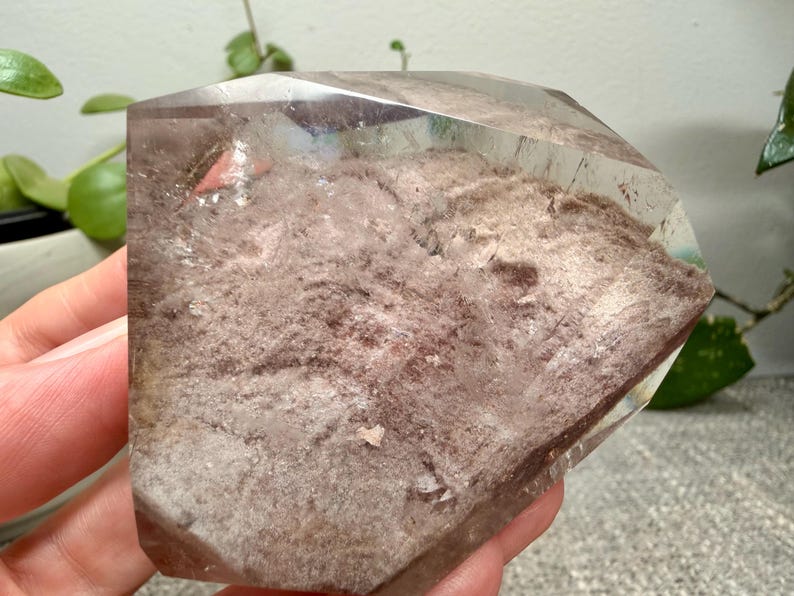 May include: A polished, faceted quartz crystal specimen featuring a clear, transparent outer layer and a textured, dusty rose-colored interior inclusion. The stone has a flat, geometric top surface and a wide, irregular base with visible mineral deposits.