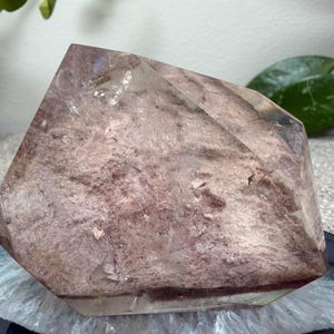 May include: A polished crystal point featuring a translucent, angular shape with a dusty rose and mauve interior inclusion. The stone displays a mix of clear, glass-like edges and a textured, earthy core with deep reddish-purple horizontal banding.