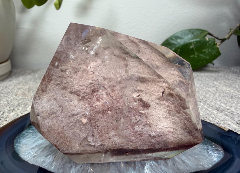 May include: A polished crystal point featuring a translucent, angular shape with a dusty rose and mauve interior inclusion. The stone displays a mix of clear, glass-like edges and a textured, earthy core with deep reddish-purple horizontal banding.