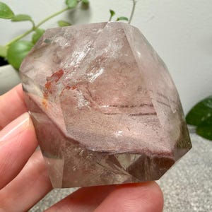 May include: A polished, multifaceted quartz crystal featuring a clear, transparent exterior with internal inclusions of dusty rose and reddish-brown mineral deposits. The stone has a geometric, angular shape with distinct flat facets and smooth edges.