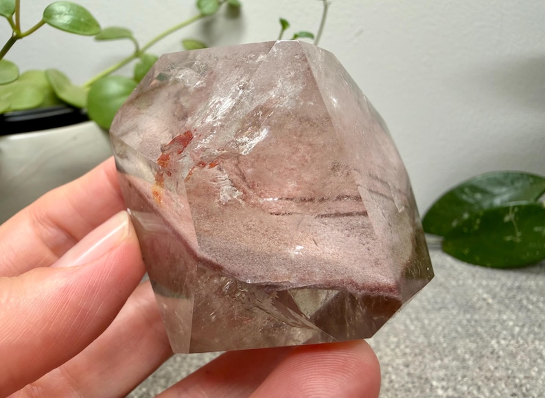 May include: A polished, multifaceted quartz crystal featuring a clear, transparent exterior with internal inclusions of dusty rose and reddish-brown mineral deposits. The stone has a geometric, angular shape with distinct flat facets and smooth edges.