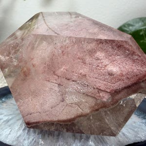 May include: A polished, multifaceted quartz crystal specimen featuring a prominent, textured reddish-pink inclusion layer within its clear, transparent body. The stone has a geometric, angular shape with smooth, reflective surfaces and visible internal fractures.