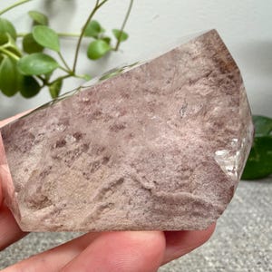 May include: A polished, semi-translucent crystal specimen featuring a dusty rose and mauve interior with a textured, mottled appearance. The stone has a smooth, angled top surface and a clear, crystalline structure visible along the edges.