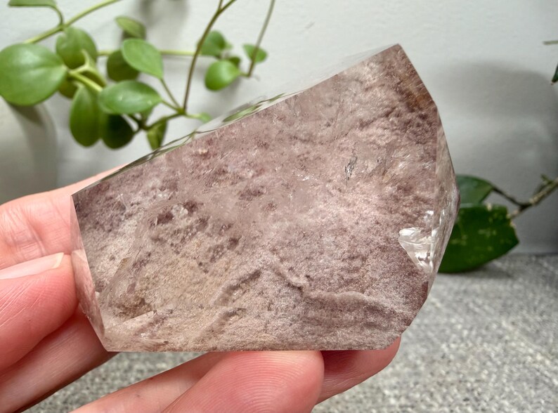 May include: A polished, semi-translucent crystal specimen featuring a dusty rose and mauve interior with a textured, mottled appearance. The stone has a smooth, angled top surface and a clear, crystalline structure visible along the edges.