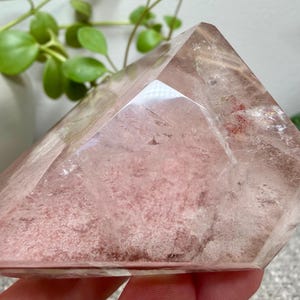 May include: A polished, faceted quartz crystal point with a clear, transparent exterior and a dense, dusty rose-colored mineral inclusion concentrated within the center. The stone features sharp, geometric edges and a smooth, reflective surface.