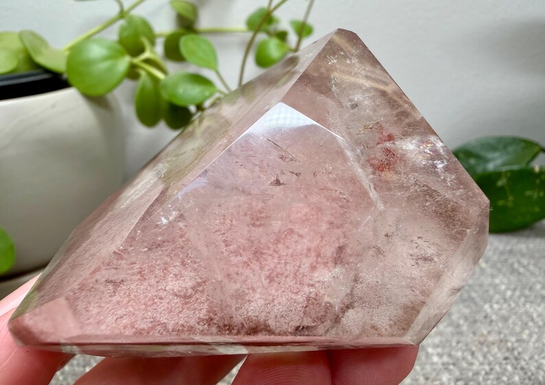 May include: A polished, faceted quartz crystal point with a clear, transparent exterior and a dense, dusty rose-colored mineral inclusion concentrated within the center. The stone features sharp, geometric edges and a smooth, reflective surface.
