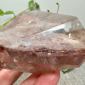 May include: A polished, multifaceted quartz crystal featuring a prominent, textured internal inclusion layer with earthy brown and reddish-pink hues. The crystal has a clear, transparent outer shell with sharp, geometric edges and a smooth surface.