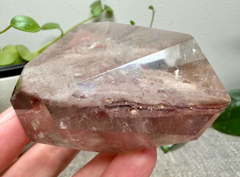 May include: A polished, multifaceted quartz crystal featuring a prominent, textured internal inclusion layer with earthy brown and reddish-pink hues. The crystal has a clear, transparent outer shell with sharp, geometric edges and a smooth surface.