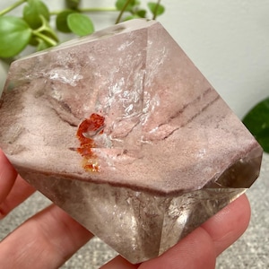 May include: A faceted, translucent quartz crystal featuring a prominent phantom inclusion with dusty pink and brown horizontal banding. A small, vibrant orange mineral deposit is visible near the center of the clear, geometric crystal structure.