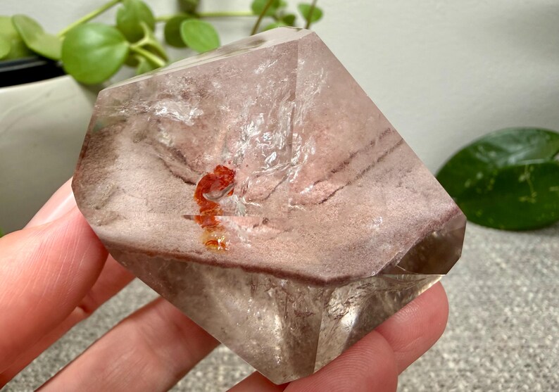 May include: A faceted, translucent quartz crystal featuring a prominent phantom inclusion with dusty pink and brown horizontal banding. A small, vibrant orange mineral deposit is visible near the center of the clear, geometric crystal structure.