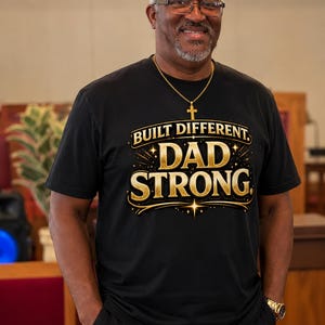 Op de afbeelding: Een zwart t-shirt met gouden letters met de tekst "BUILT DIFFERENT. DAD STRONG." Het shirt wordt gedragen door een man met een bril en een gouden kruis ketting. Hij glimlacht en heeft zijn handen in zijn zakken.