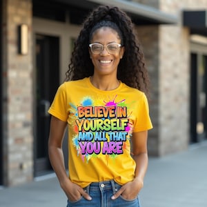 Puede incluir: Una camiseta amarilla con el texto "BELIEVE IN YOURSELF AND ALL THAT YOU ARE" en un diseño colorido estilo grafiti. La camiseta tiene mangas cortas y cuello redondo.