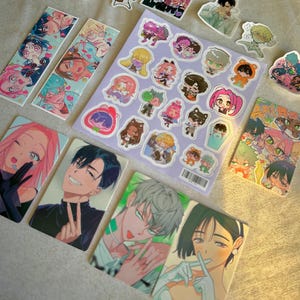 Könnte beinhalten: Eine Sammlung von Aufklebern, Lesezeichen und Karten im Anime-Stil mit farbenfrohen Illustrationen verschiedener Charaktere. Die Aufkleber befinden sich auf einem lila Bogen, und die Lesezeichen und Karten haben individuelle Charakterdesigns.