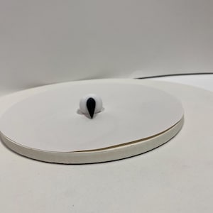May include: A small, white, rounded object with a black, teardrop-shaped protrusion. The object is centered on a white, circular platform with a slightly raised edge, set against a plain white background. The object appears to be a decorative element.