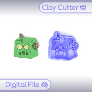 May include: A green clay cutter and a blue clay cutter, both shaped like a cartoon monster. The green cutter has a brown and tan knife and a red and white target. The blue cutter is a mold of the green cutter. The text "Clay Cutter" and "Digital File" are visible.