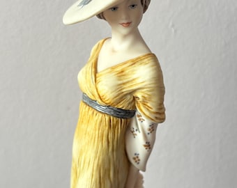 Könnte beinhalten: Eine Porzellanfigur einer Frau in einem gelben Kleid und einem weißen Hut mit blauem Band. Sie hält einen weißen Sonnenschirm in ihrer rechten Hand.
