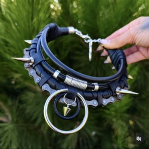Może przedstawiać: Czarny naszyjnik typu choker z metalowym łańcuchem rowerowym, skóropodobnym owinięciem i srebrnymi kolcami. Projekt zawiera duży okrągły wisiorek z mniejszym wewnętrznym pierścieniem oraz wiszącą zawieszkę w kształcie kolca, zapinany na metalowy zatrzask.