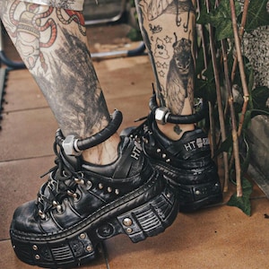 Puede incluir: Zapatos de plataforma negros con detalles metálicos, tachuelas y cordones. Los zapatos tienen una suela gruesa y una caña alta. El tobillo está adornado con una banda de cuero negro y una tuerca de metal plateado. El texto "HT 87" está en el lateral.