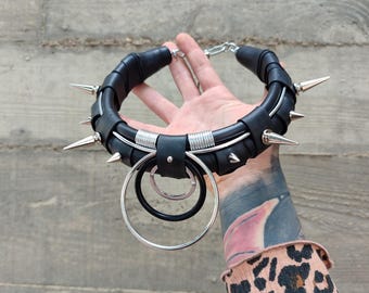 May include: A black leather choker necklace with silver-colored metal accents. The choker features multiple silver spikes, two silver rings, and a chain clasp. The necklace is held up against a gray concrete background.
