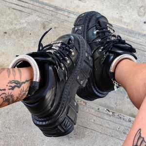 May include: A pair of black platform shoes with thick lug soles, black laces, and silver-toned metal hardware. Each shoe features a metallic silver coiled spring anklet accessory wrapped around the ankle area, creating a distinct industrial look.