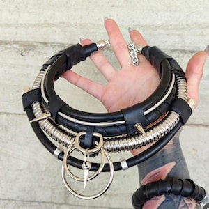 Può includere: Una collana girocollo in pelle nera e metallo argentato con un design a strati. La collana presenta una serie di tubi neri, spirali in metallo argentato e accenti a punta. Due anelli argentati e un ciondolo pendono dalla parte anteriore.