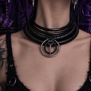 Peut inclure: Un collier ras-du-cou noir à plusieurs épaisseurs composé de bandes lisses et striées, centré par un grand anneau en métal argenté et un anneau noir plus petit, agrémenté d'un pendentif pointe en métal argenté suspendu à une boucle en cuir.