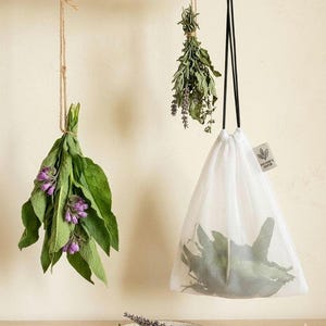 May include: A white mesh drawstring bag containing green leaves, hanging next to a bundle of green leaves with purple flowers and a bundle of dried herbs. A small tag on the bag features a leaf icon and the text "WETSIE'S DOTON".